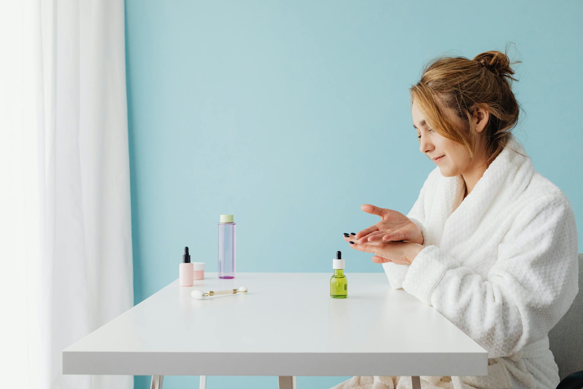 woman in a bathrobe sitting by a table and applying a product to her hands